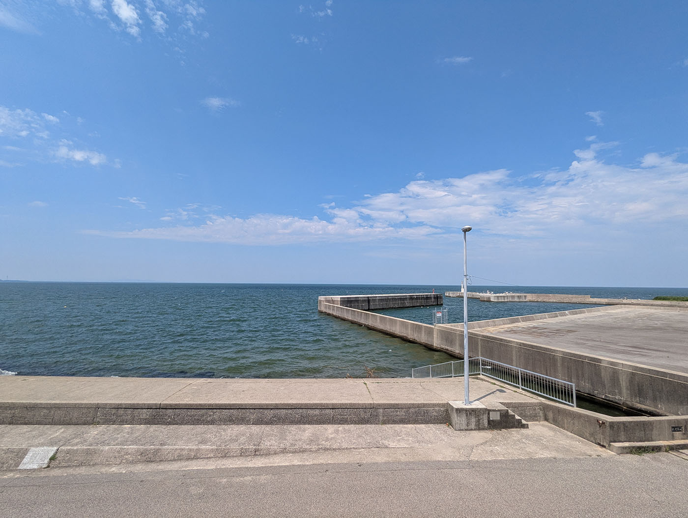 道の駅ウェーブパークなめりかわから見える富山湾の防波堤と港。コンクリートの岸壁が海に突き出し、青い海が水平線まで続く。夏の空には白い雲が浮かぶ。