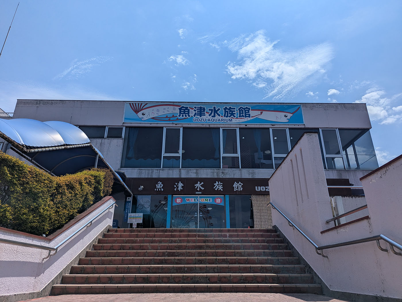 魚津水族館の正面入口。階段を上った先に「魚津水族館 UOZU AQUARIUM」の看板が掲げられ、「WELCOME」の文字が来館者を迎える。昭和の面影を残すレトロな建物で、青い空と夏雲が広がる。