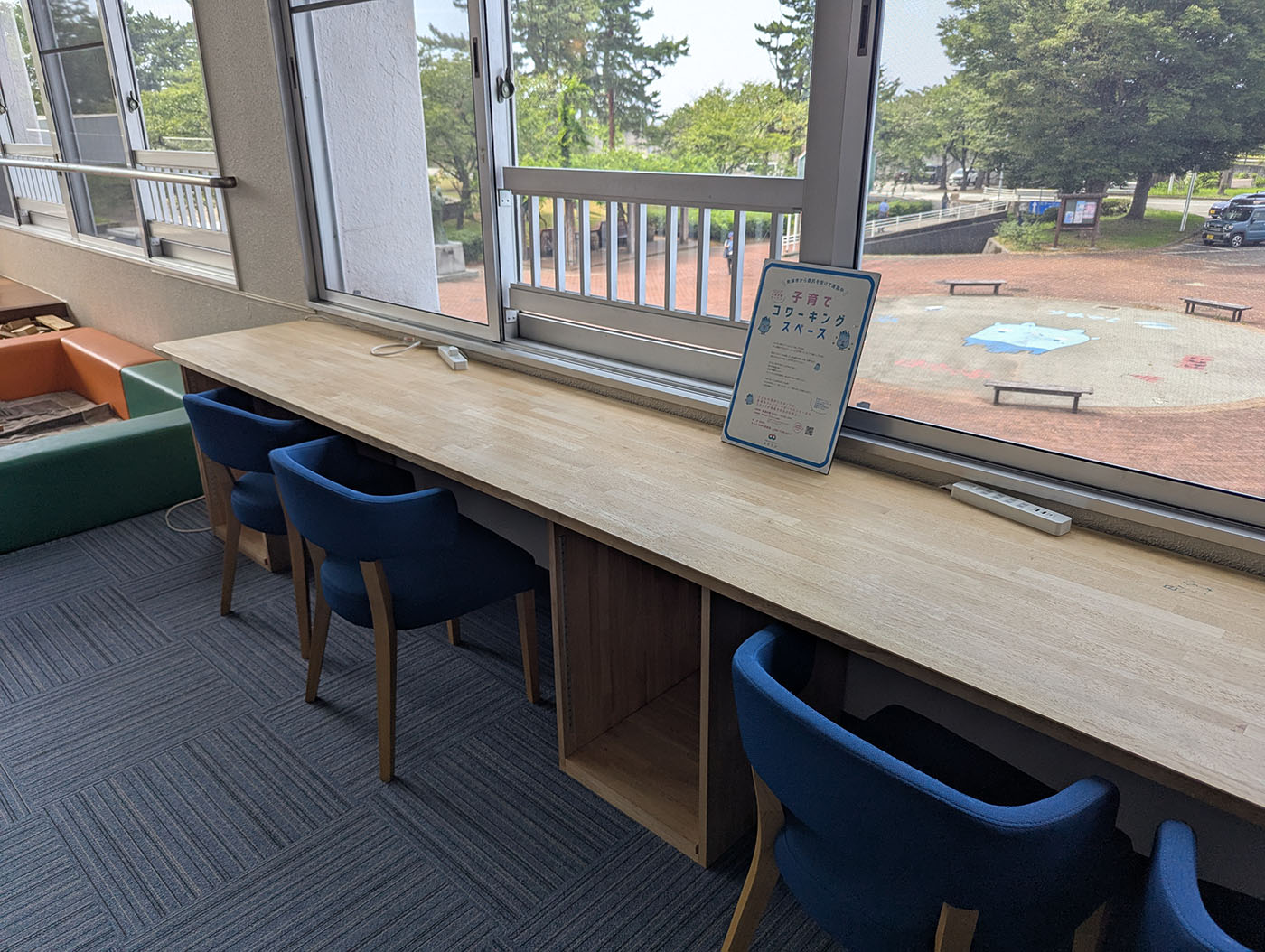 魚津水族館隣接の無料休憩所にある子育てコワーキングスペース。窓際に長いカウンターデスクが設置され、青い椅子が3脚並んでいる。デスクには「子育てコワーキングスペース」の案内板が置かれ、窓の外には魚津総合公園の遊び場が見える。フリーWi-Fiと電源完備。