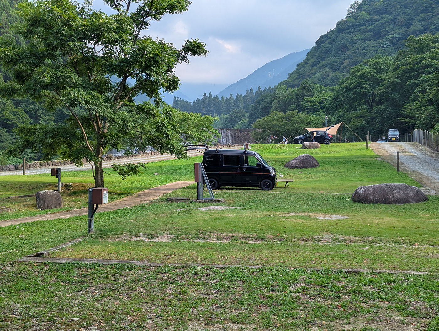 片貝山ノ守キャンプ場の広々とした芝生サイトに停められた黒いハイゼットカーゴ。背景には山々と木々が連なり、静かな夏の午後の空気が漂う。