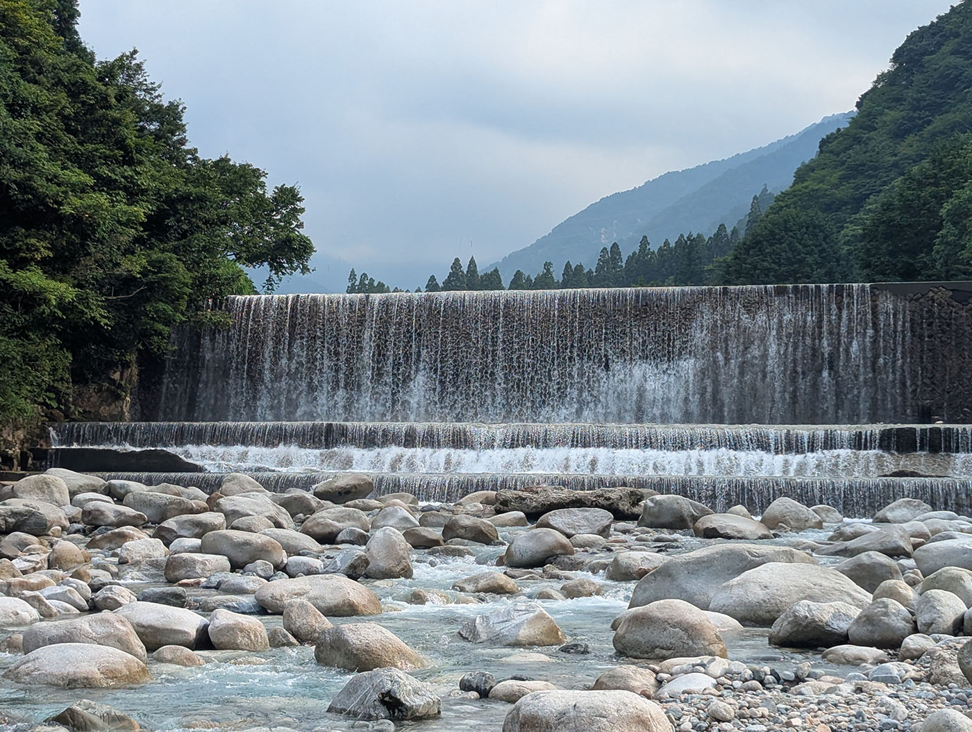 片貝川上流の砂防ダムから流れ落ちる水。3段の階段状のダムから白い水が滝のように流れ落ち、下流の河原には無数の丸い石が転がる。両岸は深い緑の山々に囲まれ、霧がかった山の稜線が遠くに見える。