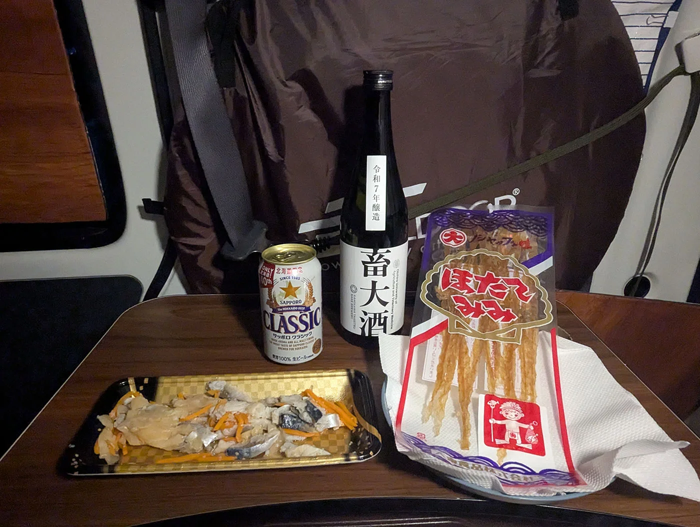 車中泊仕様の軽バン車内で、簡易テーブルに並べたサッポロクラシック、畜大酒、氷下魚の干物と惣菜。冬の十勝で開店した一人居酒屋車中泊の夜。
