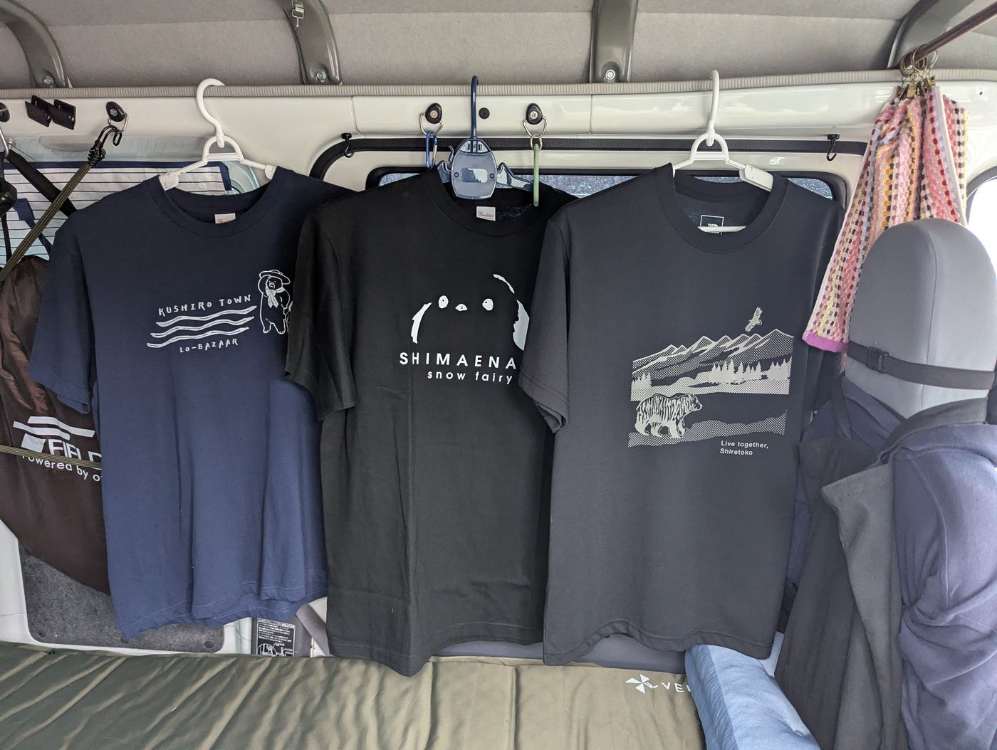 車内にハンガーで掛けた三枚の北海道Tシャツ。釧路タウン、シマエナガ、知床のデザインが並んでいる