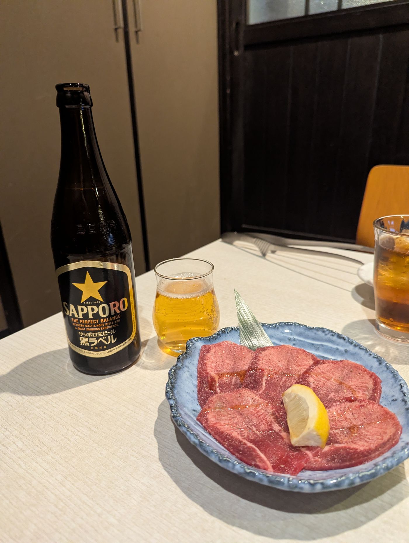 帯広の焼肉店で、サッポロ黒ラベルと小さなグラス、レモンを添えたタンの皿がテーブルに並ぶ静かな夜の食卓
