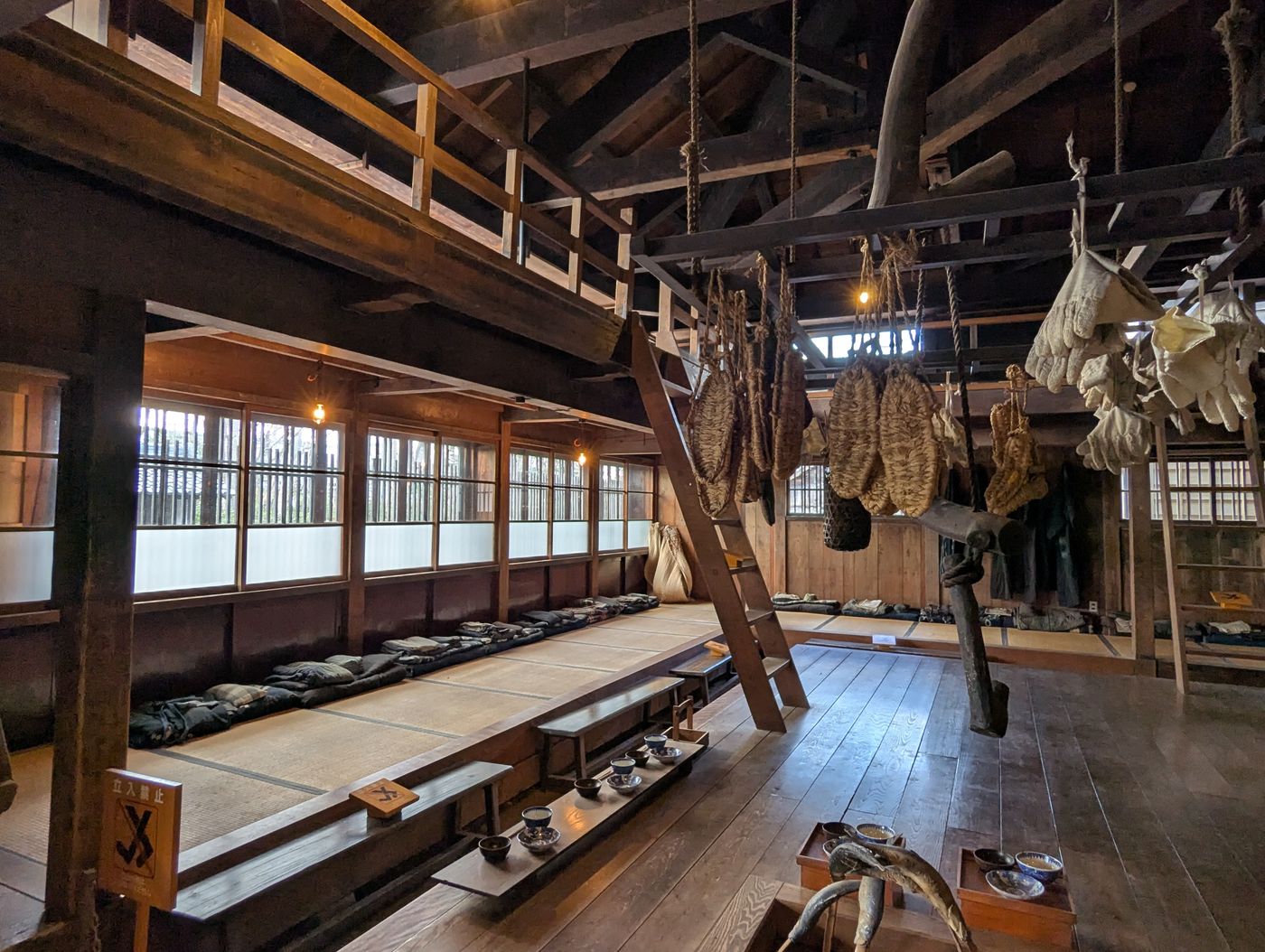 囲炉裏のある漁場建築の内部で、梁から網や道具が吊るされ、畳敷きの雑魚寝スペースが並ぶ様子