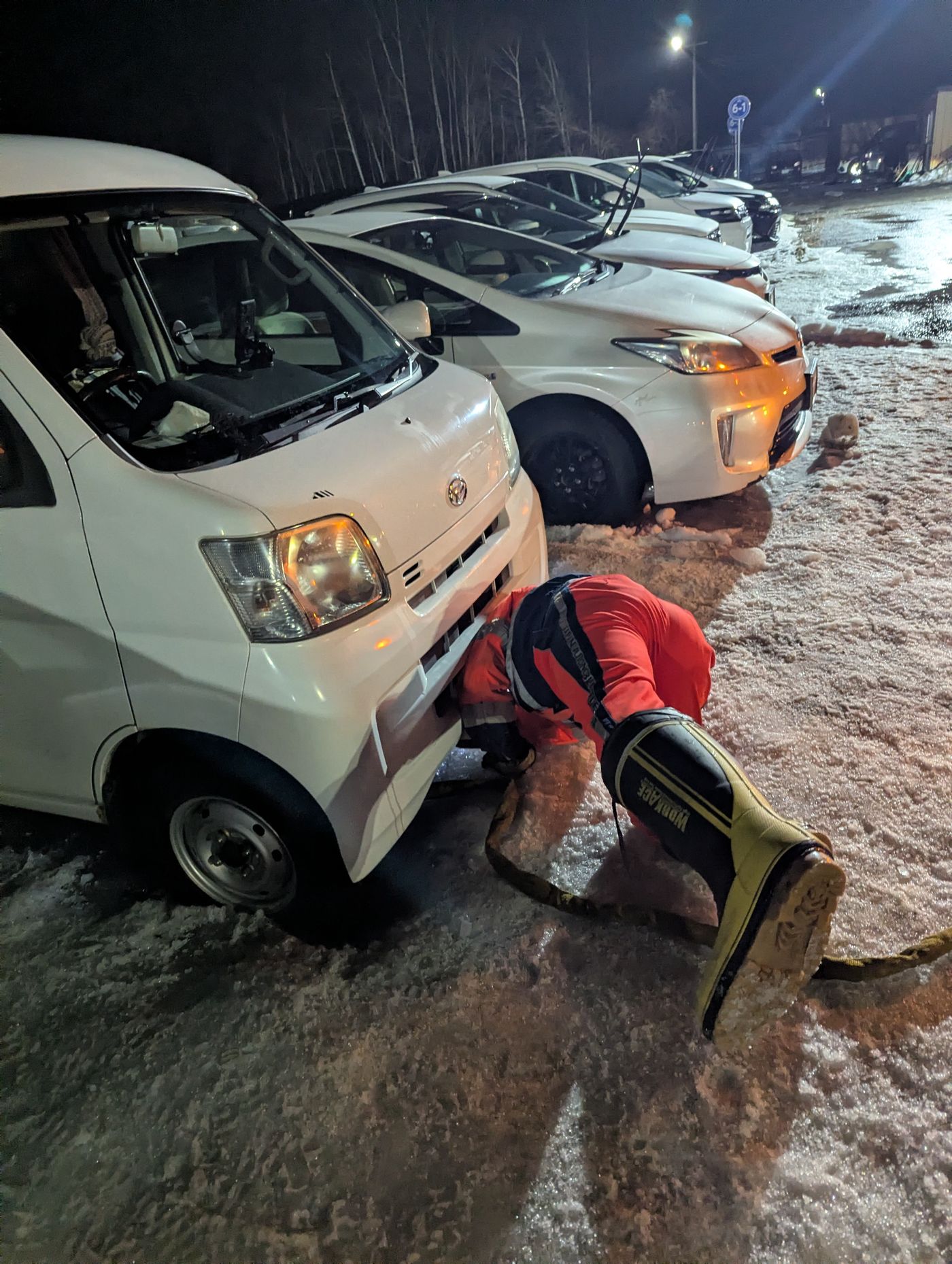 夜の雪に覆われた駐車場で、JAFの作業員が白い軽バンの下にもぐり込み、牽引ロープを取り付けている様子