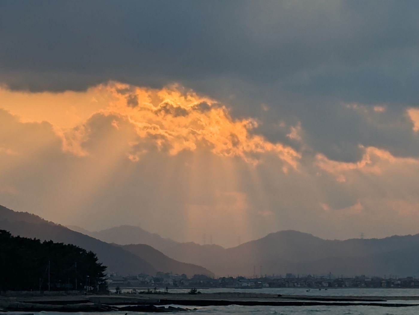若狭和田ビーチから望む夕暮れ。燃えるような橙色の雲が灰色の空に広がり、山々のシルエットと海面が静かに広がる。
